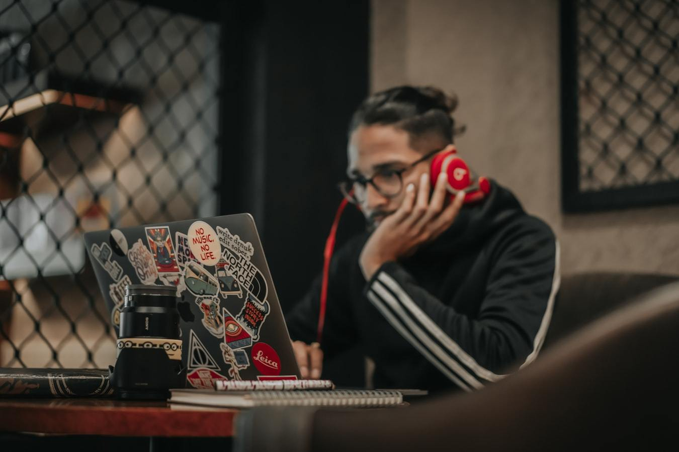 Person wearing a black hoody and red headphones works at a laptop with lots of arty stickers